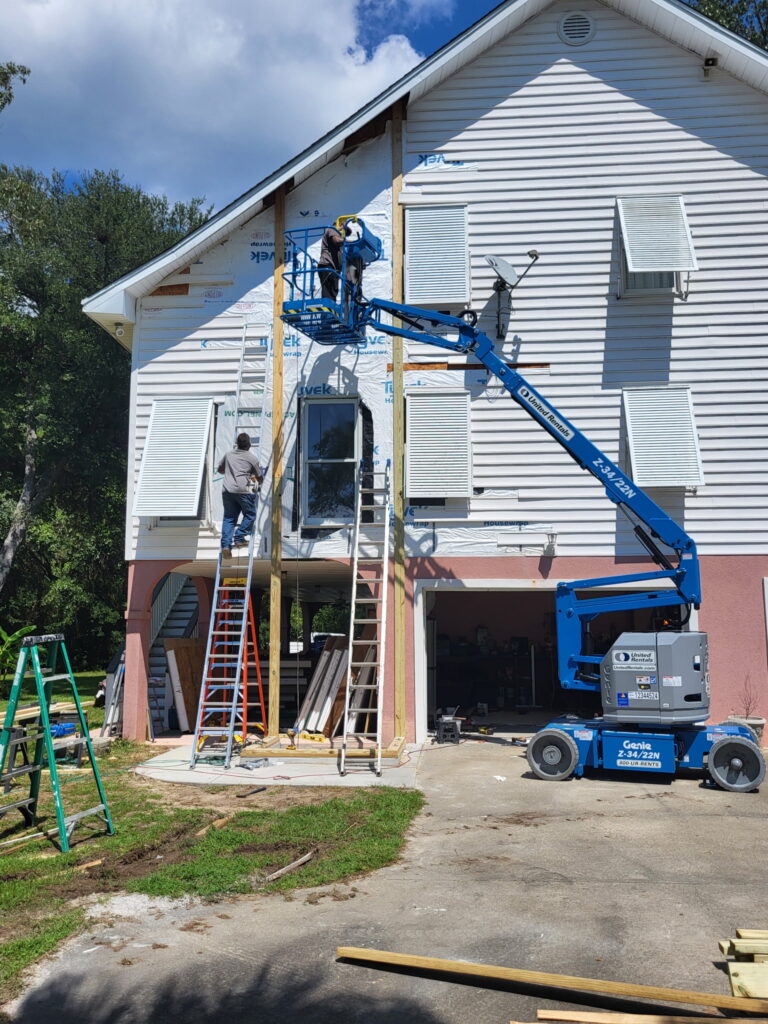A crew member on a lift installs framing near the roofline of the Meggett home as the SIP elevator shaft structure rises alongside the existing gable roof and house wrap.