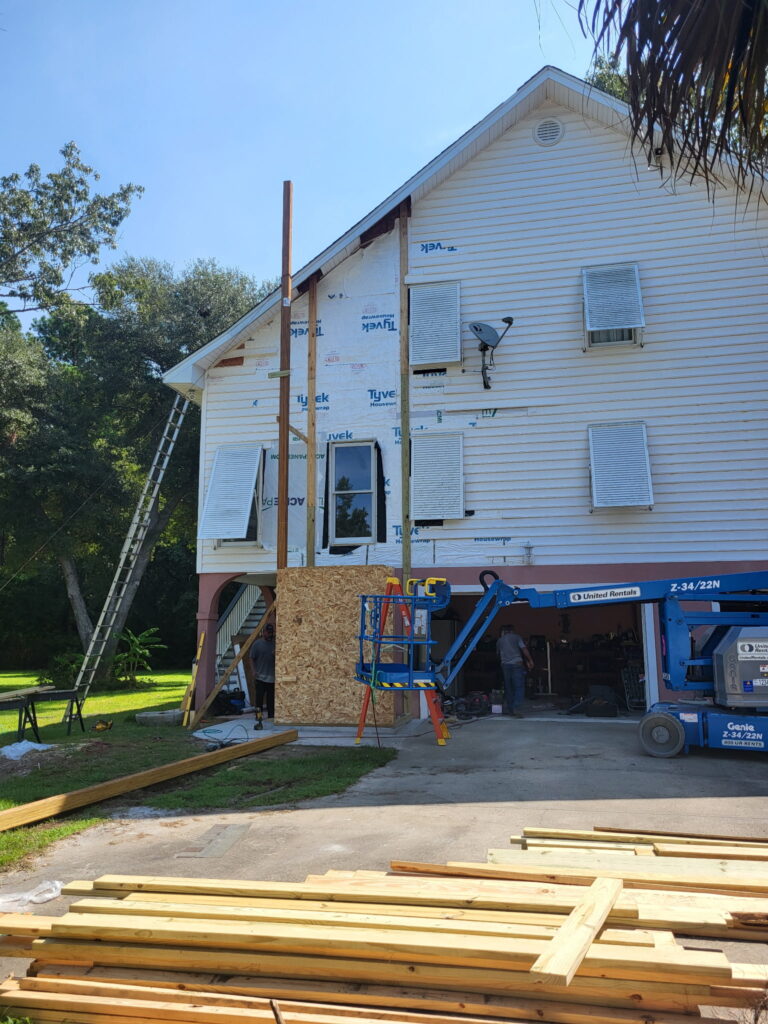 Two crew members work from a boom lift to frame the SIP elevator shaft at the gable end of the home, with OSB sheathing and engineered wood columns visible against the existing siding.