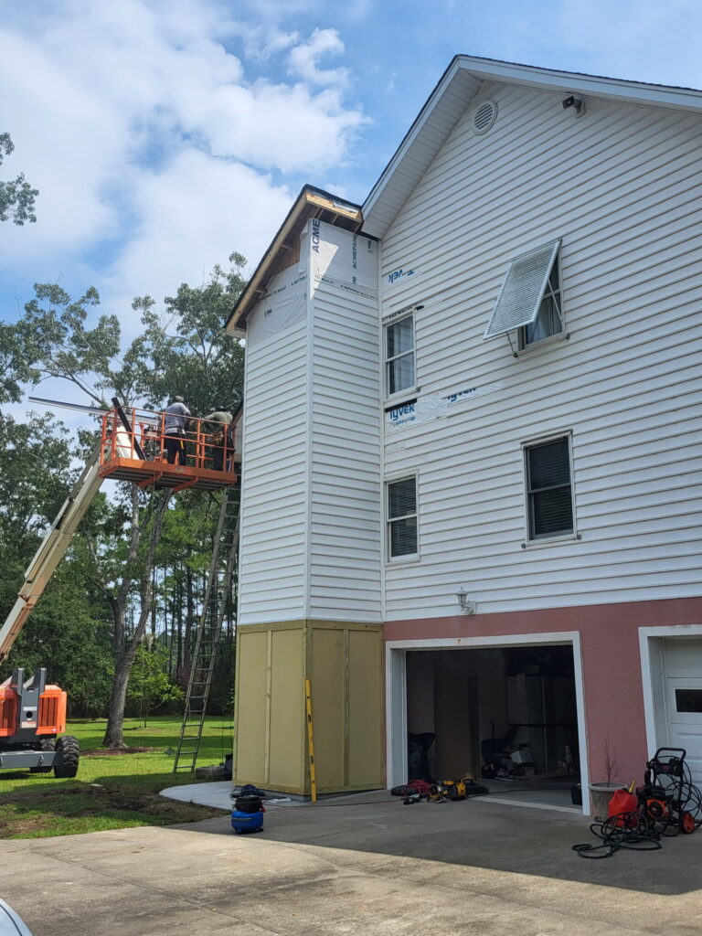 A crew member works from a raised boom lift near the roofline of the three-story SIP elevator shaft addition on the Meggett, South Carolina home, with sheathing installed down to the garage level.